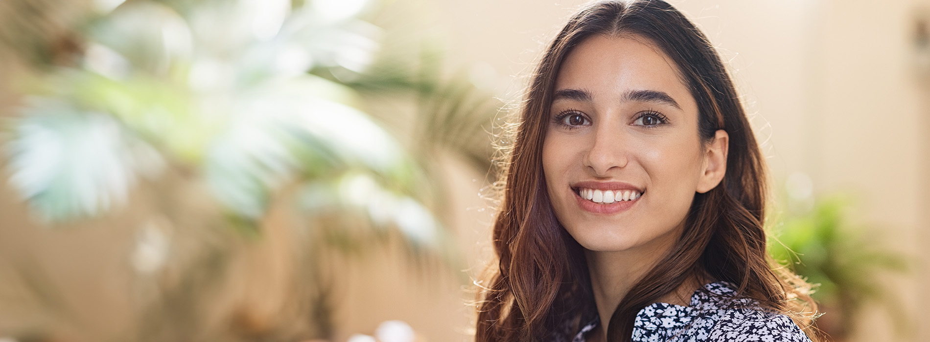 A woman with dark hair smiling at the camera, standing in front of a potted plant.